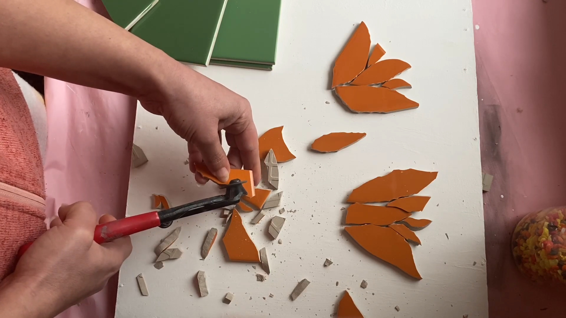 A person uses tile nippers to shape orange ceramic pieces into petal-like forms for a tulip mosaic design on a white surface, with green tiles and a jar of mosaic pieces nearby