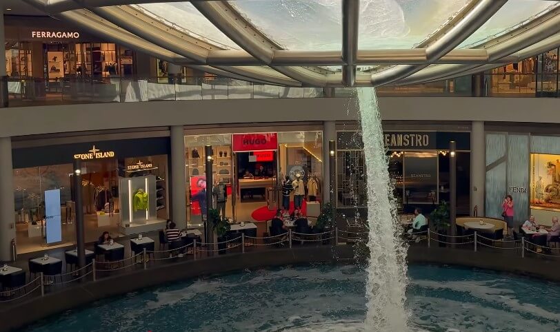 An indoor waterfall cascades near a collection of luxury retail stores