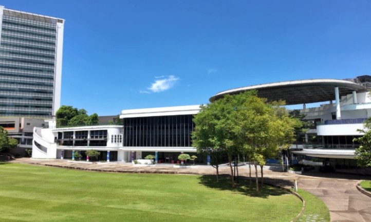 Modern buildings with large glass windows and a distinctive curved roof sit beside a lush green lawn and trees under a bright, clear blue sky
