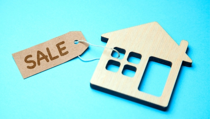 Wooden house model with a sale tag on a blue background, representing US property purchases by foreign buyers.