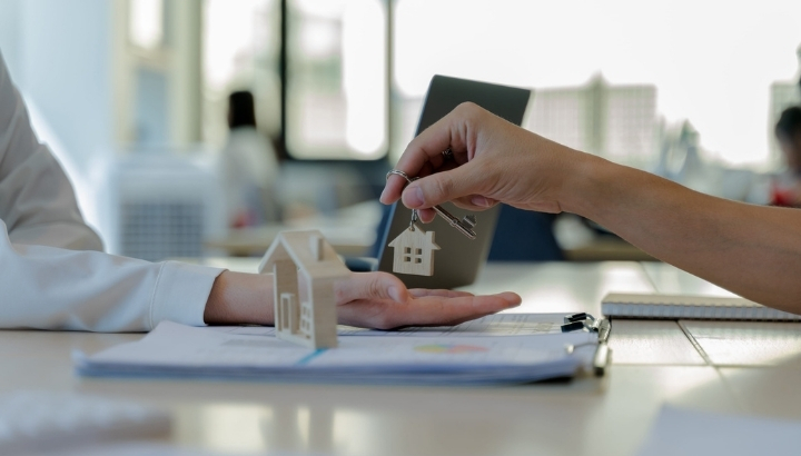 Real estate agent handing house keys to a buyer across a desk with a miniature house model